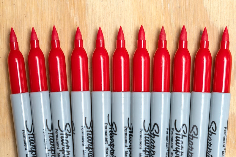 May include: A row of ten red Sharpie permanent markers lined up on a wooden surface.
