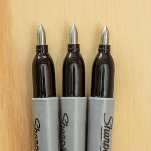 K&ouml;nnte beinhalten: Drei Sharpie Permanentmarker mit silbernen Spitzen und schwarzen Sch&auml;ften. Die Marker haben graue Geh&auml;use mit dem aufgedruckten Sharpie-Logo. Der Hintergrund ist eine hellbraune Holzoberfl&auml;che.