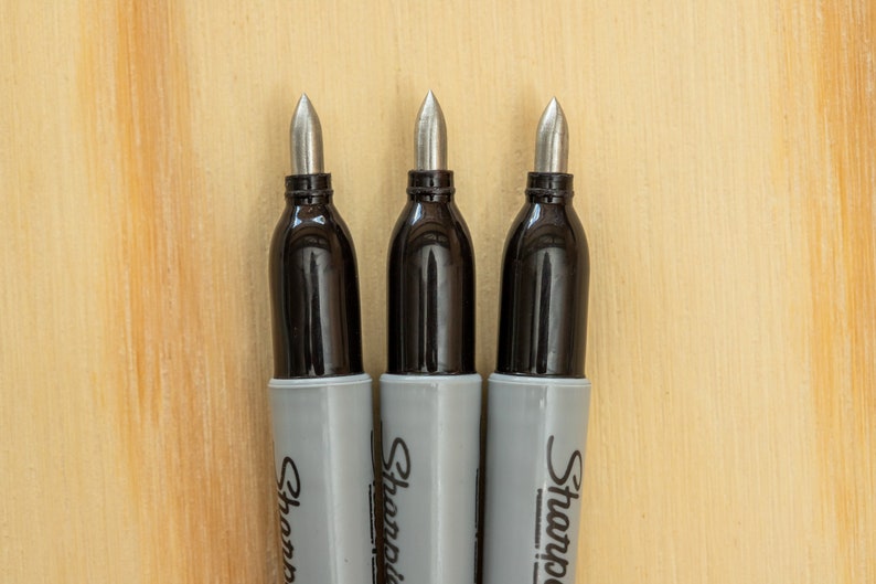 K&ouml;nnte beinhalten: Drei Sharpie Permanentmarker mit silbernen Spitzen und schwarzen Sch&auml;ften. Die Marker haben graue Geh&auml;use mit dem aufgedruckten Sharpie-Logo. Der Hintergrund ist eine hellbraune Holzoberfl&auml;che.