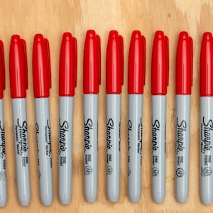 May include: A row of 13 red Sharpie fine point permanent markers on a wood surface. The markers are all gray with black lettering.