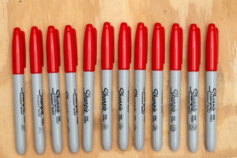 May include: A row of 13 red Sharpie fine point permanent markers on a wood surface. The markers are all gray with black lettering.
