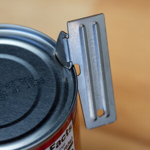 May include: A silver can opener with a black handle is being used to open a can of food. The can has a black lid with a printed date code.