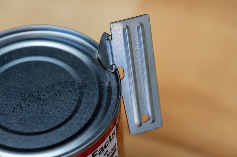 May include: A silver can opener with a black handle is being used to open a can of food. The can has a black lid with a printed date code.