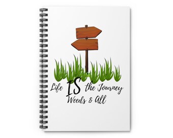 May include: A white spiral-bound notebook featuring a graphic of two brown wooden directional signs standing in green grass. Below the illustration, the black text reads: Life IS the Journey Weeds & All. The notebook has a black metal coil spine.