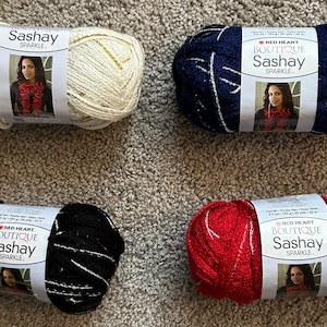 May include: Four skeins of Red Heart Boutique Sashay Sparkle yarn in white with gold sparkle, navy blue with silver sparkle, black with silver sparkle, and red with silver sparkle.