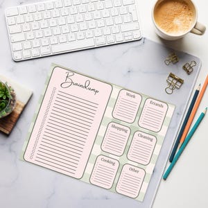 May include: A pink and green Braindump notepad with sections for lists. The notepad has a lined section for notes and smaller sections labeled "Work", "Errands", "Shopping", "Cleaning", "Cooking", and "Other". A white keyboard and coffee cup are in the background.