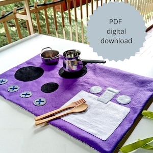 May include: A purple felt play kitchen set with a stove, pots, utensils, and a placemat. The stove has black burner circles and blue cross-shaped knobs. The set includes a small pot, a saucepan, a fork, and a spoon.