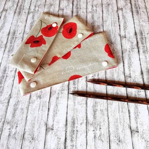 Puede incluir: Tres estuches de agujas de tela con estampado de flores de amapola y botones blancos. Dos agujas de tejer de madera están sobre una superficie de madera blanca.