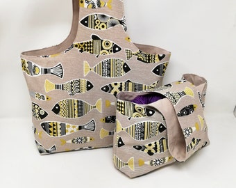 May include: Two beige fabric tote bags with a black, white, and yellow fish pattern. The larger bag has a single handle, while the smaller bag has two handles. The smaller bag is filled with a purple ball of yarn.