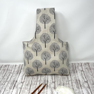 Op de afbeelding: Een beige stoffen tas met een herhalend zwart boompjespatroon en een enkele geïntegreerde handgreep. De tas heeft een gestructureerde vorm met een platte bodem en een hoge, gecentreerde handgreep, voorzien van een print van gestileerde bomen.