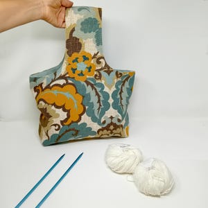 May include: A floral patterned tote bag in shades of teal, gold, and brown is held by a hand. Two balls of white yarn and two blue knitting needles are on a white surface. The bag has a handle and a rectangular shape.