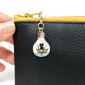 Può includere: Un ciondolo dorato con un gatto nero in una barca all'interno di una lampadina. Il ciondolo è attaccato a una zip nera.