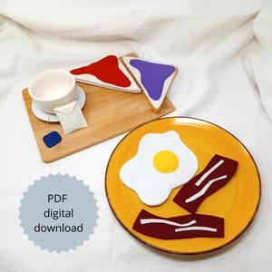May include: A felt breakfast set with a yellow plate holding a fried egg and bacon strips. A wooden tray holds toast with red and purple spreads, a teacup, and a tea bag. The image includes the text "PDF digital download."