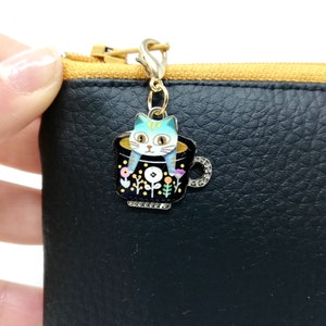 Può includere: Un ciondolo dorato con una tazza nera e bianca con un gatto blu seduto all'interno. La tazza ha un bordo bianco ed è decorata con fiori gialli e rosa. Il ciondolo è attaccato a una catena dorata.