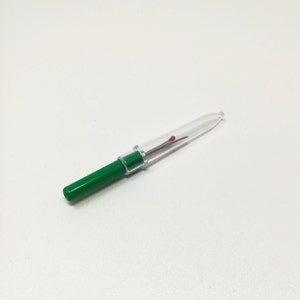 Può includere: Un tagliafilo per cucire in plastica trasparente con un tappo verde. Il tagliafilo ha un piccolo punto rosso sulla lama.
