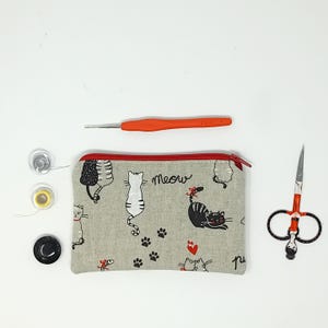 Può includere: Una collezione di forniture artigianali tra cui una pochette con cerniera grigia a tema gatto, un uncinetto arancione, piccole forbici, rocchetti di filo e un bottone nero. La pochette presenta illustrazioni di gatti e la parola "Meow".