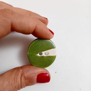 Peut inclure: Un bouton rond vert avec une bande blanche au centre et deux petits trous. Le bouton est tenu entre deux doigts sur un fond blanc. Le bouton a une surface texturée.