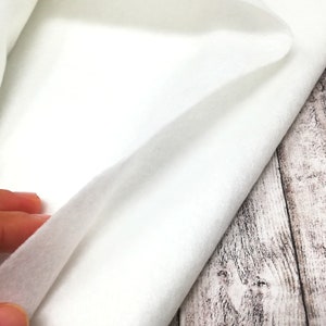 May include: White felt fabric, a soft and smooth material, folded over on itself. The fabric is shown against a light wood background.