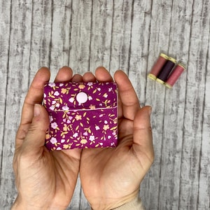 May include: A small square fabric pouch featuring a dark magenta background with a repeating pattern of tiny white and yellow flowers. The pouch is secured with a single white snap button closure. Three small spools of thread are positioned nearby.