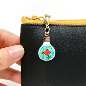 Puede incluir: Un charm dorado con forma de bombilla con un diseño pintado en azul y blanco de un pez rojo nadando en agua con burbujas. El charm está unido a un tirador de cremallera de cuero negro.