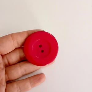 May include: A large, round, bright red button with two small holes in the center. The button is held in a hand against a white background. This sewing notion is a vibrant accent for clothing or craft projects.
