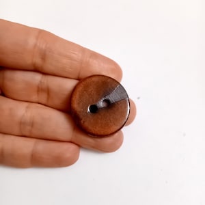 Op de afbeelding: Een ronde, bruine knoop met twee gaten en een diagonale grijze accent. De knoop heeft een glanzende afwerking, waarschijnlijk van hars. Geschikt voor naaien of knutselprojecten. Diameter ongeveer 2,5 cm.