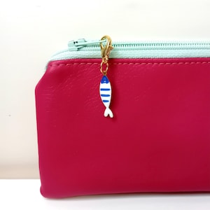 Op de afbeelding: Een roze ritszakje met een lichtblauwe rits en een gouden bedel in de vorm van een witte vis met blauwe strepen.