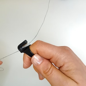 Pode incluir: Um cortador de fio de plástico preto com uma lâmina afiada, segurado na mão, cortando um fio preto fino.