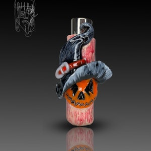 Può includere: Un accendino a tema Halloween nero e arancione con un viso di zucca e un cappello da strega.