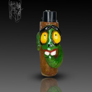 May include: A green and brown ceramic lighter with a cartoon face, wearing sunglasses and a black hat. The lighter has a green snake wrapped around it and yellow eyes.