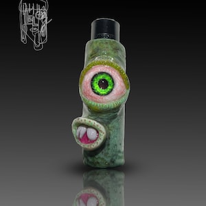 Pode incluir: Um rolha de garrafa de cerâmica verde e rosa com um olho grande e dentes. O olho é verde com uma íris rosa e os dentes são brancos.