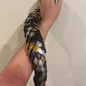 May include: A silver, gold, and black chainmail arm sleeve. The sleeve is made of interlocking metal scales.