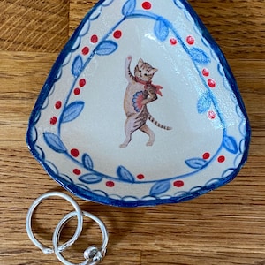 May include: A triangular ceramic trinket dish with a cream-coloured base, featuring a hand-painted illustration of a tabby cat holding a fan, surrounded by blue leaves and red berries. Two simple silver-toned metal rings rest near the dish.