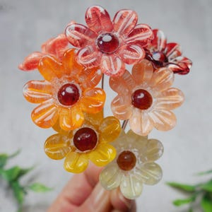 May include: A bouquet of colorful glass flowers with a daisy-like design. The flowers feature shades of orange, yellow, and red with a dark red center. The petals are translucent, and the bouquet is held together by thin metal stems.