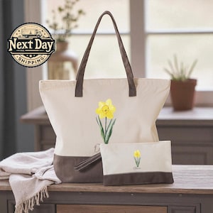 A beige tote bag with a brown handle and a small white bag with a yellow flower design are placed on a wooden surface, with a window and a plant in the background.