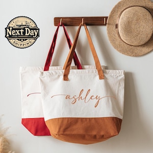May include: Two canvas tote bags with personalized text, one with a red bottom and straps, the other with a brown bottom and straps. The text "ashley" is written in a cursive font on the brown-bottomed bag. A straw hat hangs on the wall.