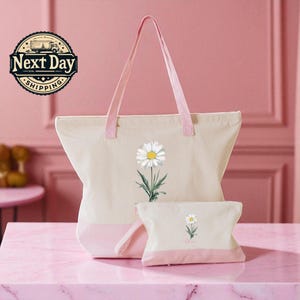 A pink tote bag with a white daisy flower design and a matching pink handle, accompanied by a smaller white bag with a similar flower design, both placed on a pink marble surface.