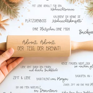 以下が含まれることがあります： テキストが書かれた木製のめん棒が手で持たれており、背景にはクリスマスの飾りがあります。めん棒には「Advent, Advent, DER TEIG, DER BRENNT！」という言葉が刻まれています。他のテキストも表示されています。