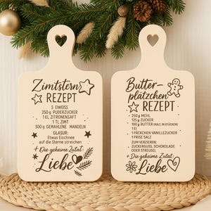 May include: Two wooden chopping boards with heart-shaped handles. One has a recipe for Zimtstern cookies, the other for Butterplätzchen cookies. Both boards feature decorative text and illustrations, ideal for festive baking.