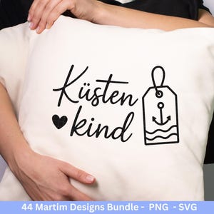 Op de afbeelding: Cr&egrave;mekleurig kussen met de Duitse woorden "K&uuml;stenkind" en een hart, plus een label met een anker en golven, allemaal in zwart. Het kussen wordt door een persoon vastgehouden.