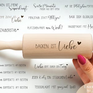 May include: A wooden rolling pin with the words "BACKEN IST Liebe" printed on it, held by a person with pink nail polish. The image includes various phrases in German related to baking, such as "Liebe geht durch den Teig" and "Keks-Zauberstab".
