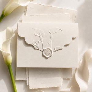 May include: An off-white envelope with a scalloped flap featuring an embossed calla lily design, sealed with a circular wax seal. The envelope rests atop matching stationery cards with deckled edges, accompanied by two white calla lily flowers.