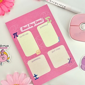 May include: Pink notepad with a geometric design and the text "Best Day Ever!"  The notepad is divided into four sections labeled "To Do List", "Schedule", "Reminders", and "Self-Care".  There are mathematical symbols like pi and sigma on the notepad.
