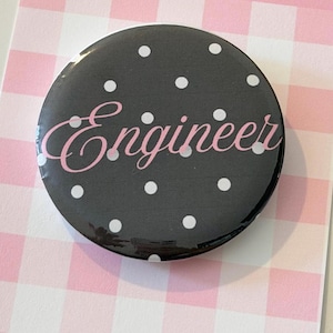 May include: A circular pin-back button featuring a black background with a white polka dot pattern. The word Engineer is printed across the center in a light pink cursive script font. The button has a glossy finish and a smooth, rounded edge.