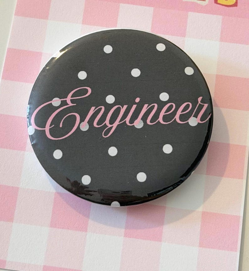 May include: A circular pin-back button featuring a black background with a white polka dot pattern. The word Engineer is printed across the center in a light pink cursive script font. The button has a glossy finish and a smooth, rounded edge.