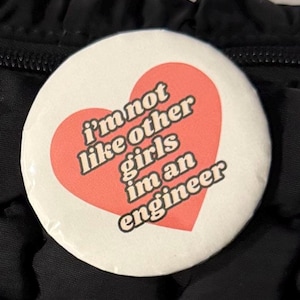 May include: A circular pin-back button featuring a white background with a large coral-colored heart in the center. The text "i'm not like other girls i'm an engineer" is printed in black, bold, sans-serif capital letters across the heart.