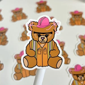 May include: A brown teddy bear sticker wearing a pink hard hat, safety glasses, and an orange vest. The bear has a heart-shaped nose and is surrounded by other bear stickers.