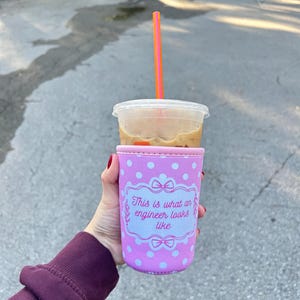 Puede incluir: Una taza de plástico transparente llena de café helado, coronada con una pajita rosa y naranja. La taza se sostiene en una funda rosa con lunares con el texto "This is what an engineer looks like."