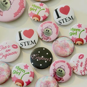 May include: A collection of round pin-back buttons featuring various designs including pink princess engineer graphics, cherry-themed stem girl illustrations, red heart I love STEM text, black polka dot engineer buttons, and colorful marbled patterns.