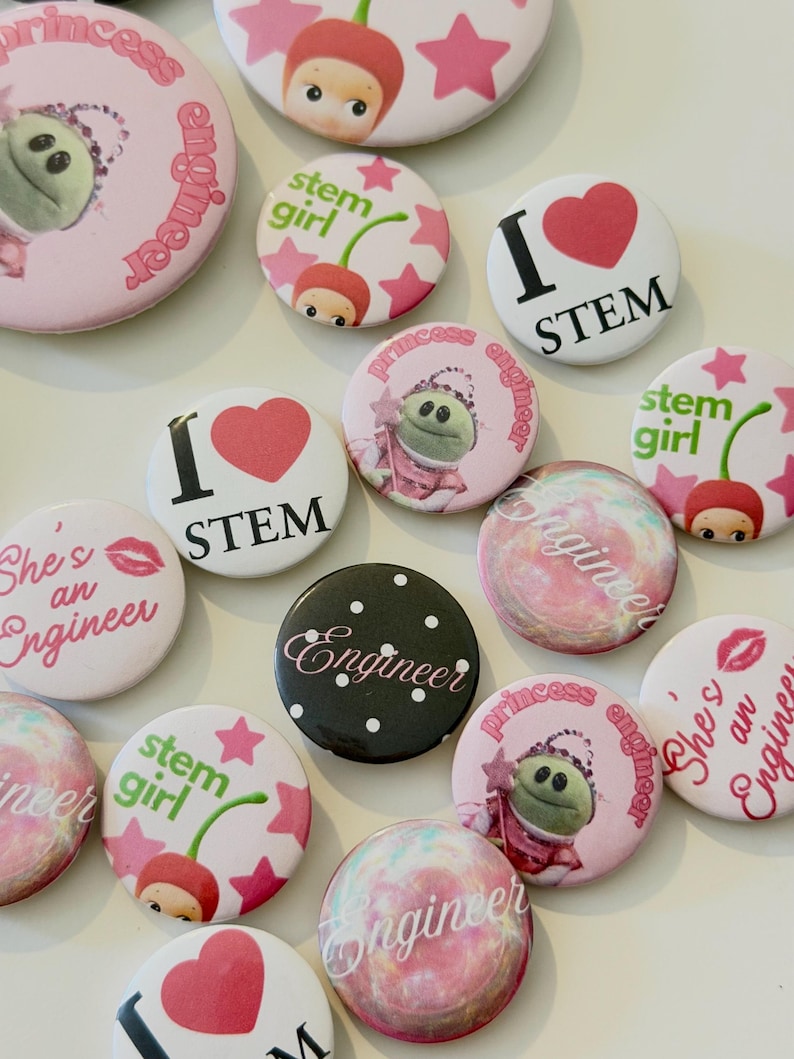 May include: A collection of round pin-back buttons featuring various designs including pink princess engineer graphics, cherry-themed stem girl illustrations, red heart I love STEM text, black polka dot engineer buttons, and colorful marbled patterns.
